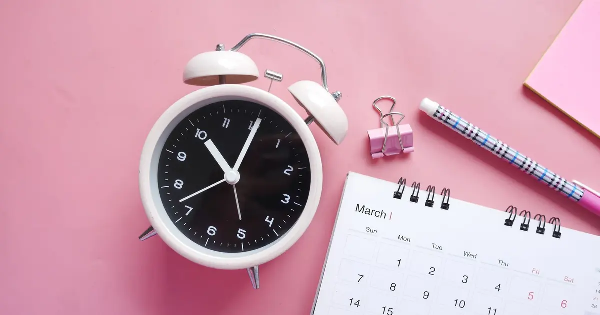 Đồng hồ báo thức và cuốn lịch trên nền hồng cùng bút và kẹp giấy